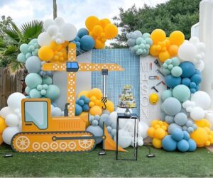 JCB-themed balloon decor with two arches, floral garland, and shimmer wall backdrop