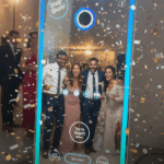 A sleek, illuminated mirror photo booth capturing a group of happy party-goers amidst falling confetti, showcasing interactive features.